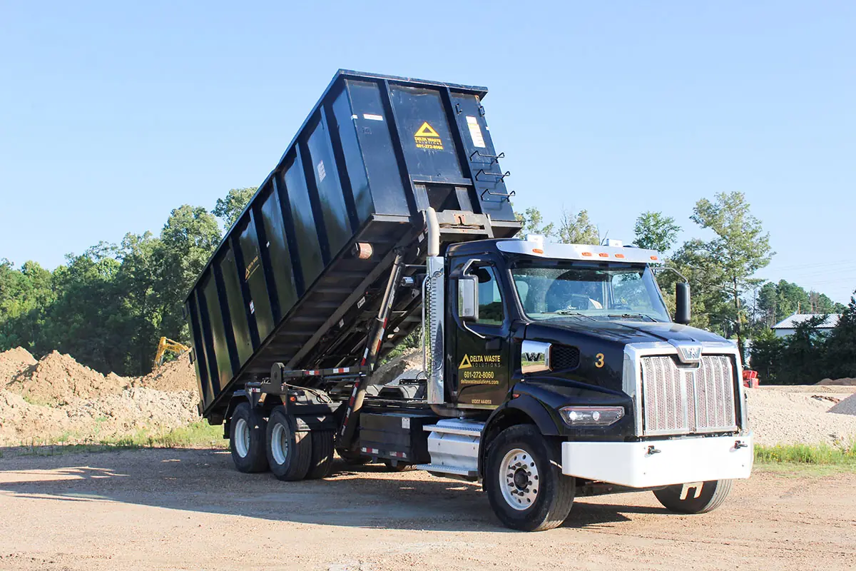Black Delta Waste Solutions hooklift truck tipping a roll-off dumpster on a sunny day, with the company logo and phone number on the door