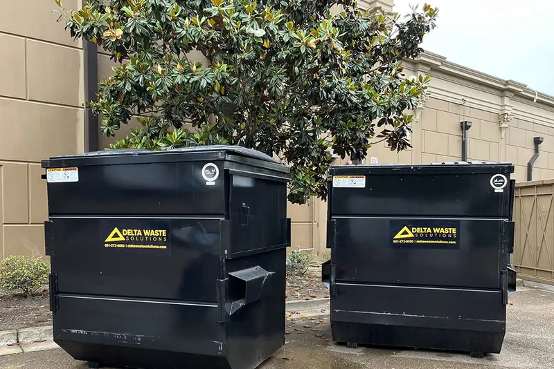 Two Delta Waste Solutions front-load dumpsters behind a commercial building